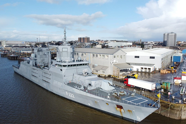 Ein Kriegsschiff, gekennzeichnet mit F 222, liegt im Hafen. Es ist ein graues Schiff mit verschiedenen Masten und Radar. Im Hintergrund sind Lagerhäuser und andere Gebäude zu sehen, während ein blauer Himmel mit einigen Wolken darüber schwebt.
