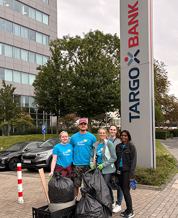 Fünf Personen stehen vor dem TARGOBANK-Gebäude. Sie tragen blaue T-Shirts und halten Müllsäcke in der Hand. Im Hintergrund sind Bäume und Autos zu sehen.