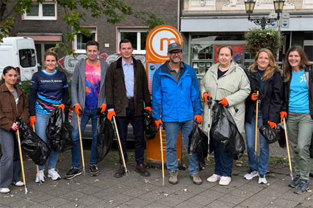 Eine Gruppe von acht Personen steht auf einem Bürgersteig, während sie Müllsäcke und Greifzangen halten. Sie tragen orangene Handschuhe und lächeln. Im Hintergrund sind einige Gebäude und ein Mülleimer sichtbar.