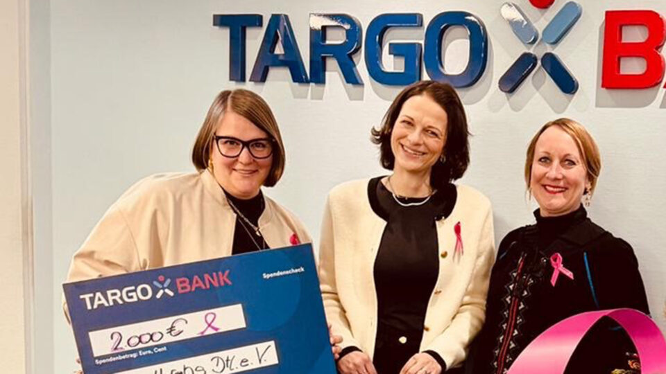Drei Frauen stehen vor dem TARGOBANK-Schild und halten einen großen Scheck über 2000 € für den Verein "Hilfe Dtl. e.V.". Die Frauen tragen jeweils ein rosa Band und lächeln.